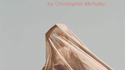 A man wearing a vest, shorts and sports shoes, balances precariously on a metal fence whilst being covered by a semi transparent mesh fabric. He is using his right arm to reach through the fabric, to try and get a better look of around him, and keep himself safe. The text - ITCH, written by Christopher McAuley - sits just above him on the fence in a bold orange colour.